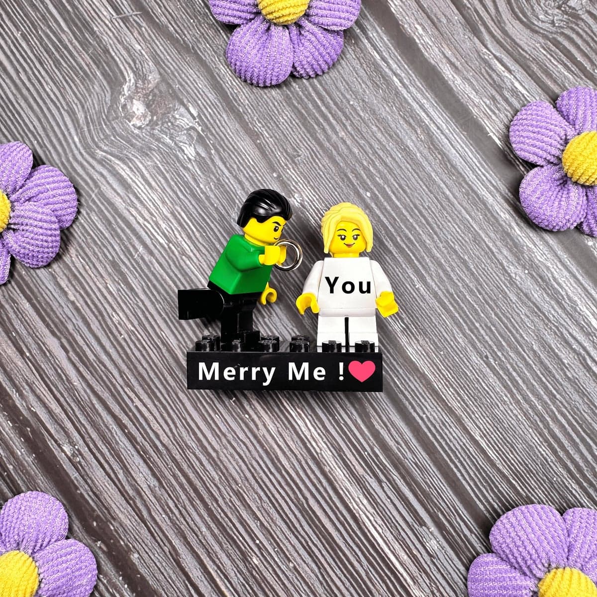 Real proposal gift example: two personalized brick figures on a black printed message brick, photographed among purple flowers